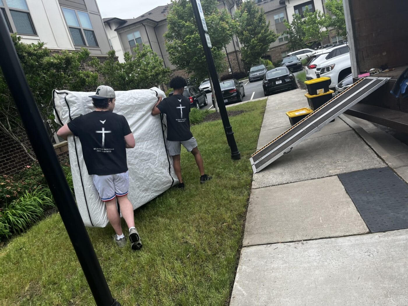 Two people are carrying a mattress toward a moving truck in a residential area, highlighting the hustle of long distance moving services. Nearby cars are parked, and several yellow-lidded bins sit on the truck's ramp, waiting to be loaded for the journey ahead.