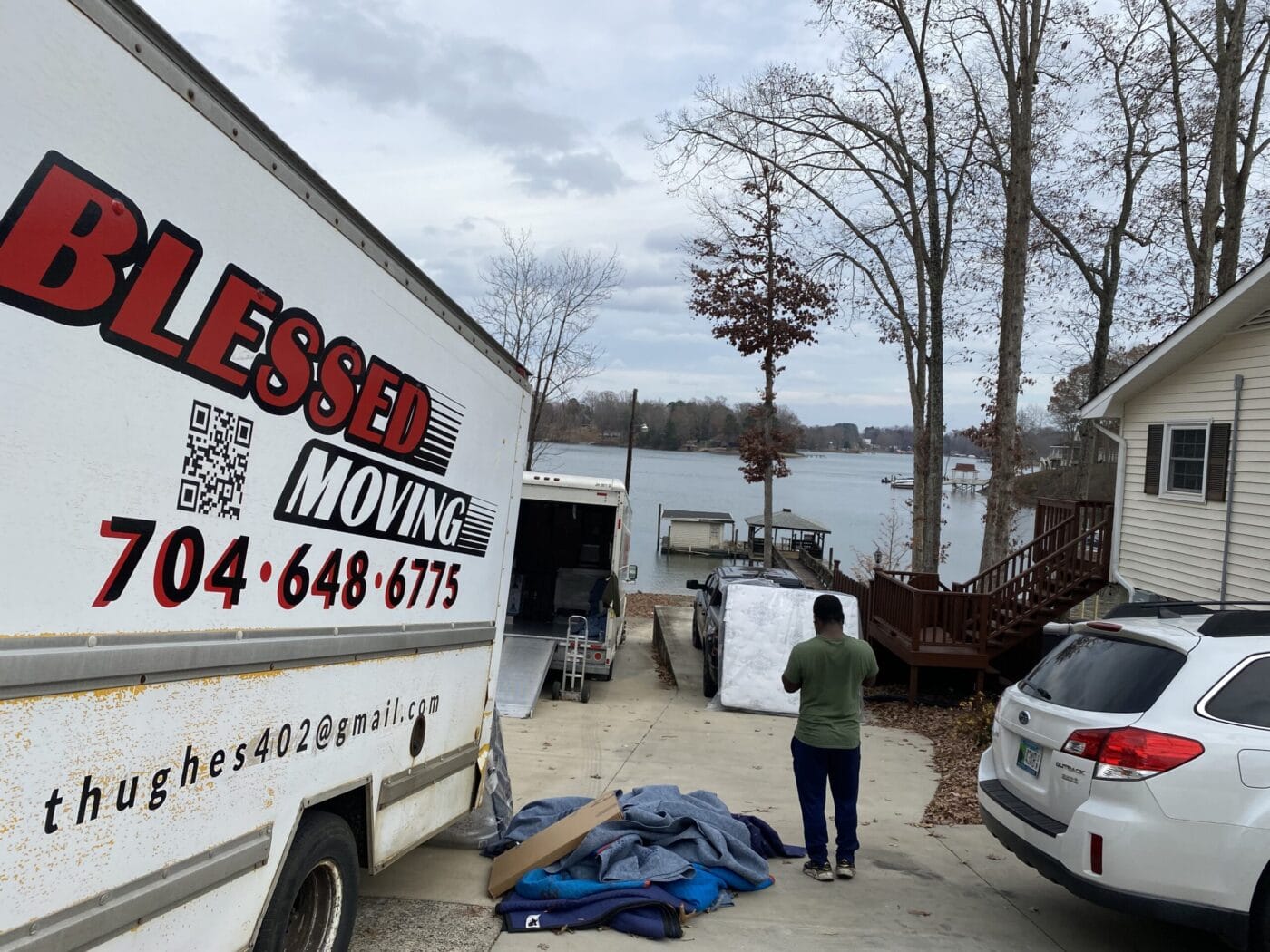 A person stands near a moving truck labeled Blessed Moving beside a lakeside house. There's furniture wrapped in blankets on the driveway and a gray SUV parked nearby.