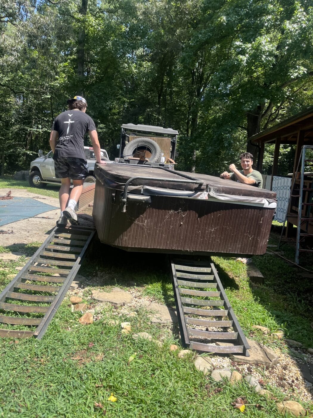 Two people, tasked with junk removal, are preparing to move a large hot tub onto a trailer using ramps in a grassy yard.