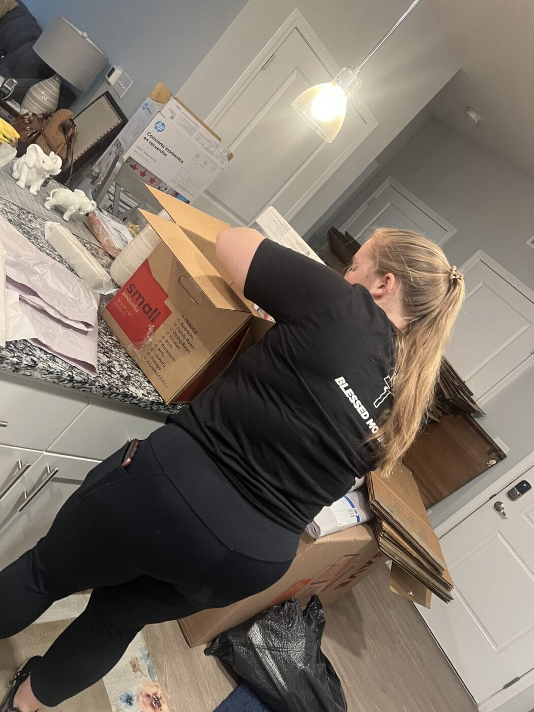 A person utilizes professional packing & unpacking services as they carefully unpack a cardboard box on a kitchen counter, surrounded by other boxes and packing materials.