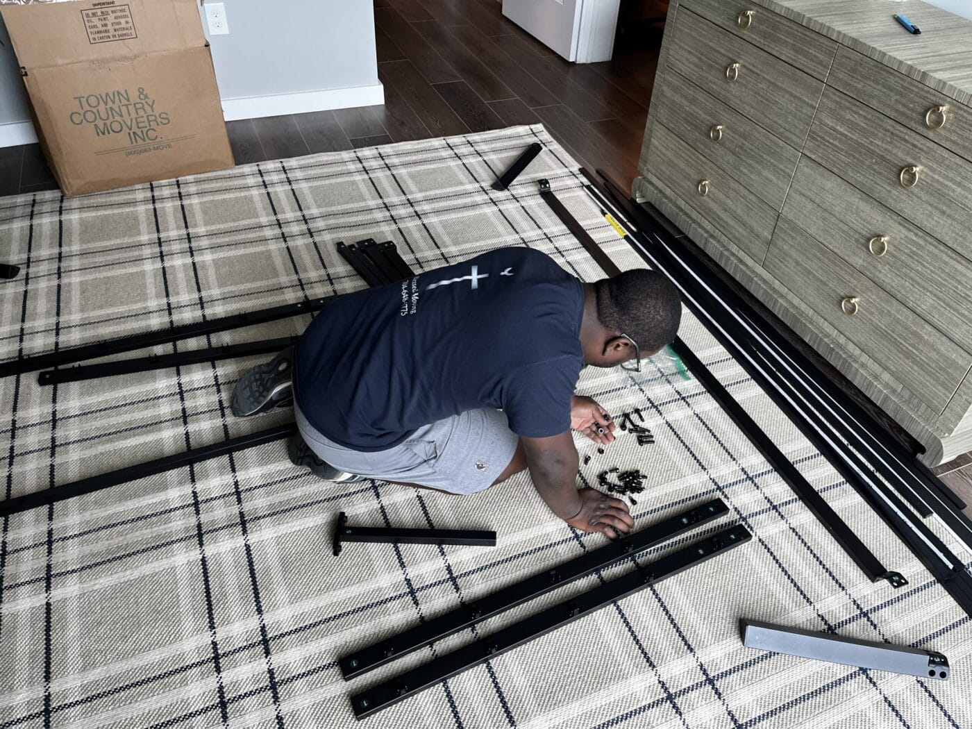 A person is assembling furniture on a plaid rug, surrounded by black metal pieces and a box labeled Town and Country Movers Inc., showcasing the efficiency of residential moving services.