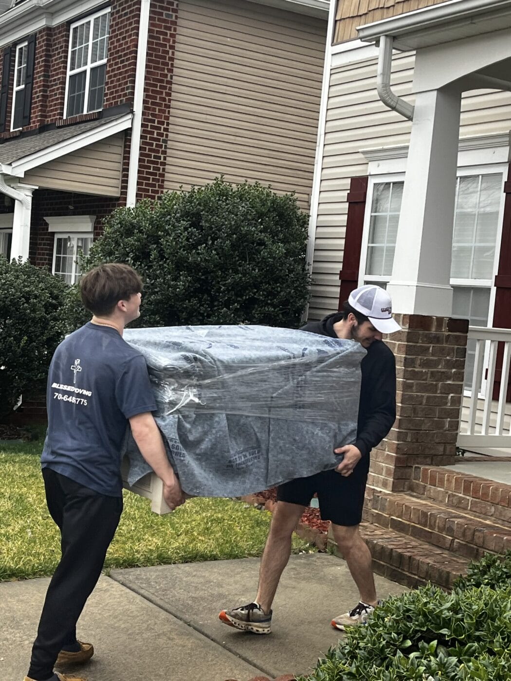 Two people from Small Moves & Deliveries are carefully carrying a large, wrapped item along a residential sidewalk, with brick steps providing a charming backdrop.