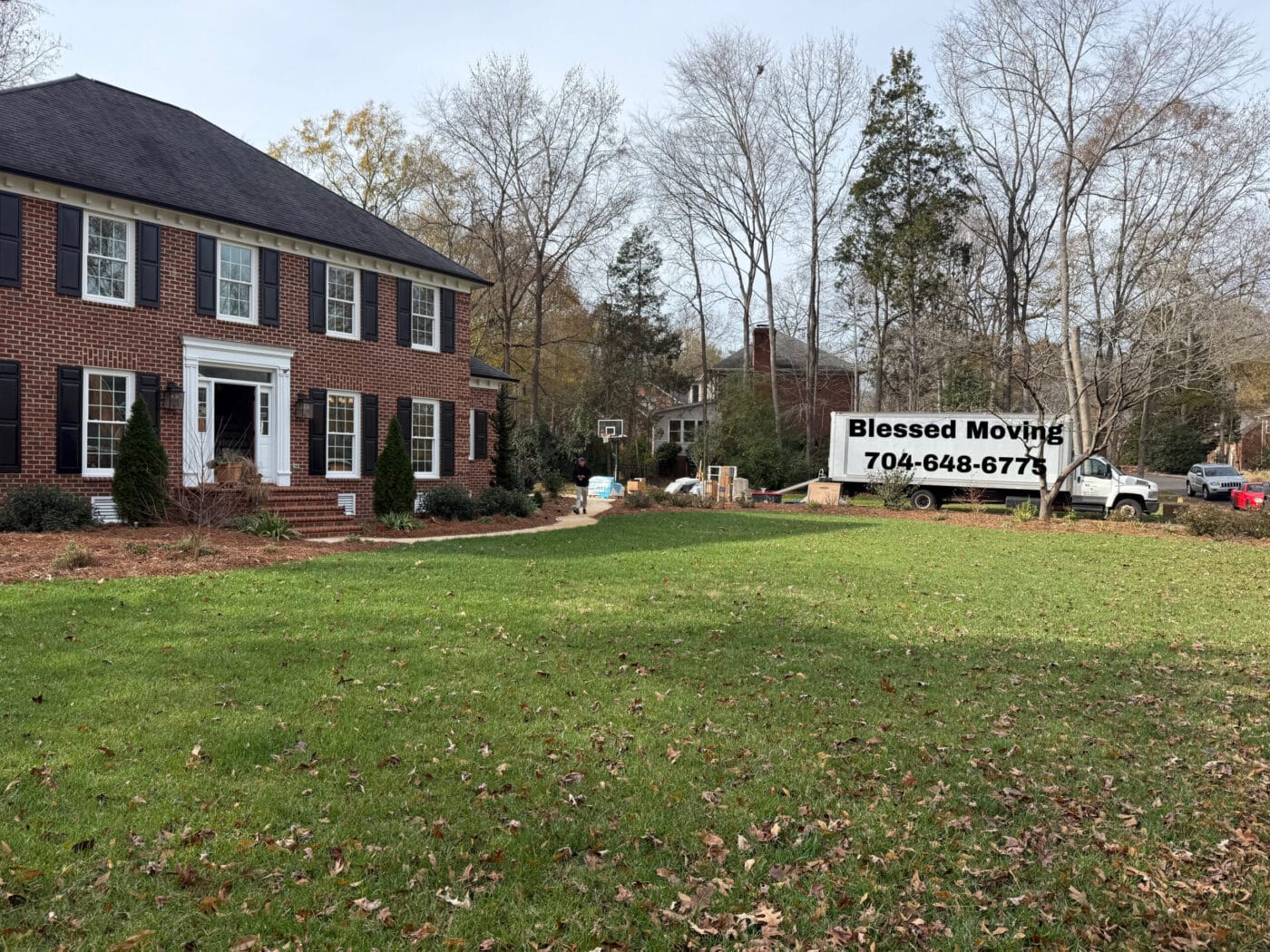 A red brick house with white columns features a lawn covered in scattered leaves. In the background, a moving truck is parked, displaying a sign for Blessed Moving with a contact number.