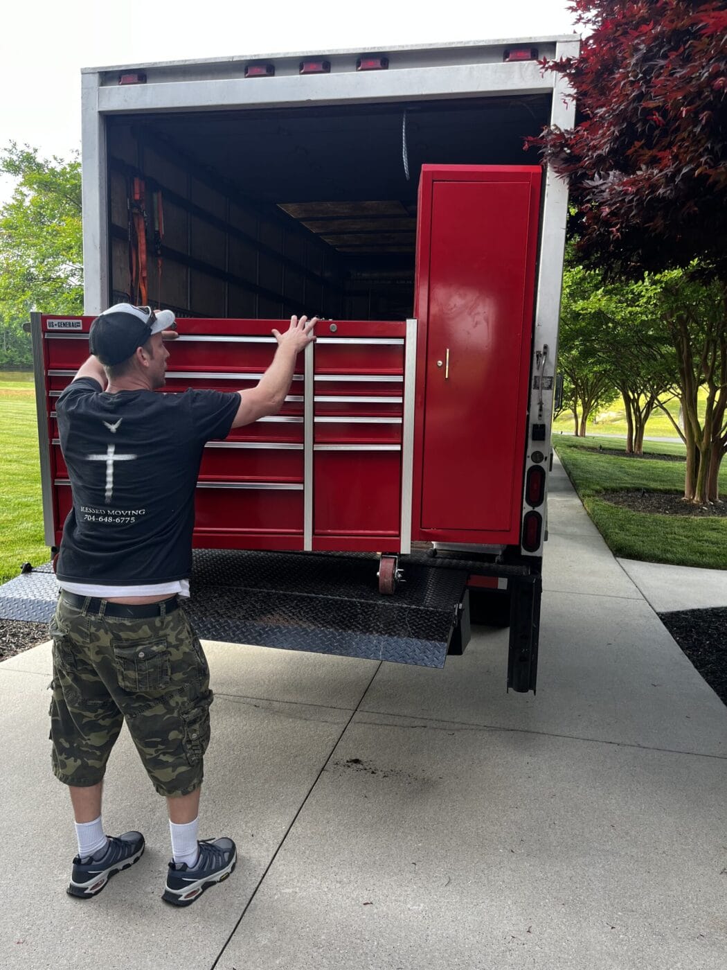 A person in camo shorts and a black shirt is loading a large red toolbox into the back of a truck on a paved driveway, reminiscent of commercial moving services. Trees and grass provide a lush backdrop.