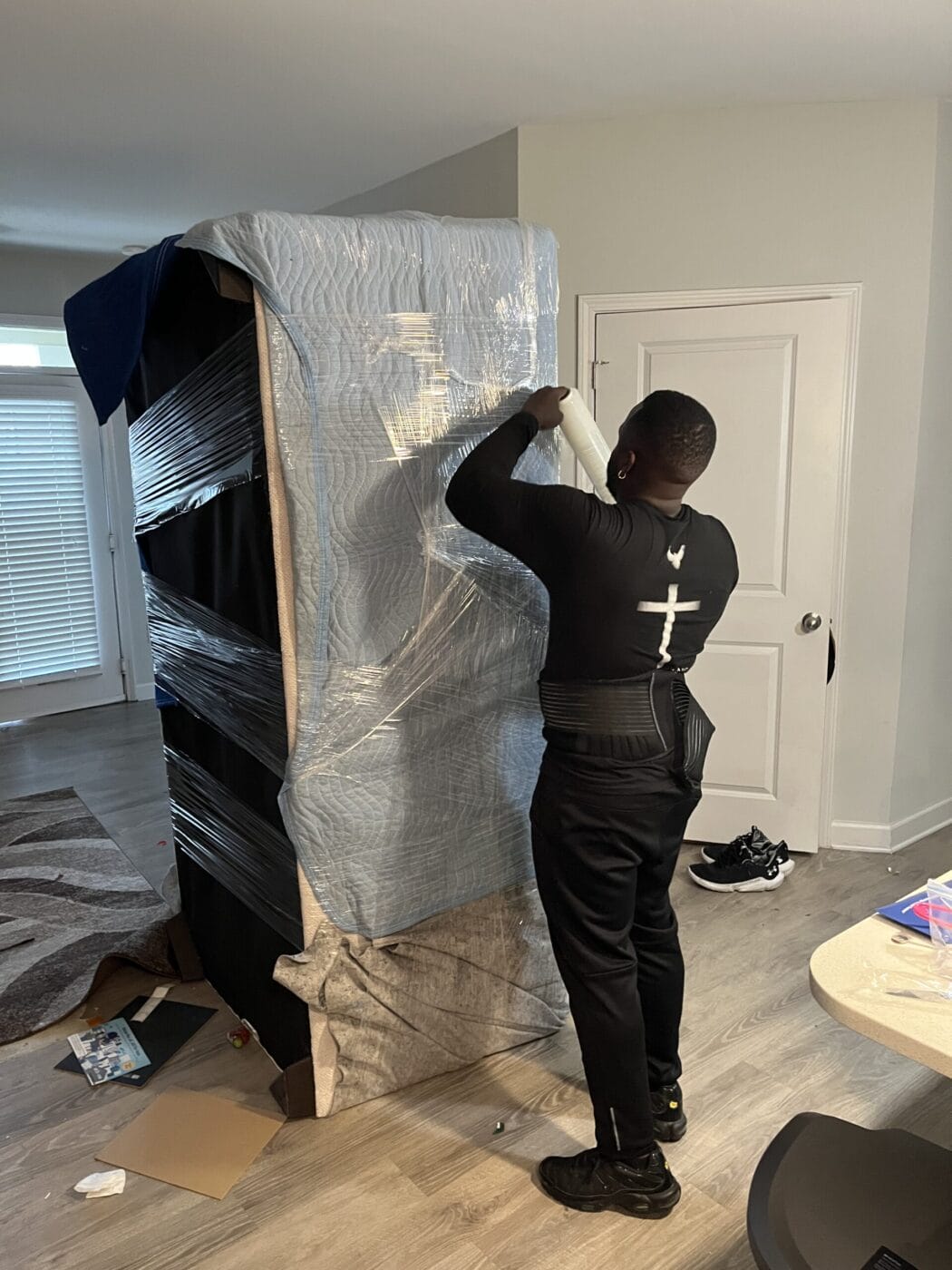 A person meticulously wraps a large, upright piece of furniture with plastic wrap amidst scattered items, showcasing the precision and care akin to White Glove Moving Services.