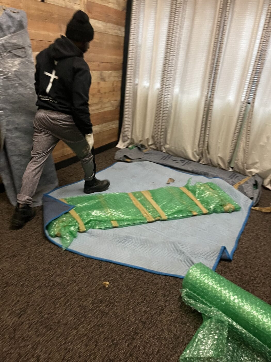 A person is walking near an object wrapped in green bubble wrap, ready for small moves and deliveries, placed on a blanket in a cozy room with wooden walls and curtains.