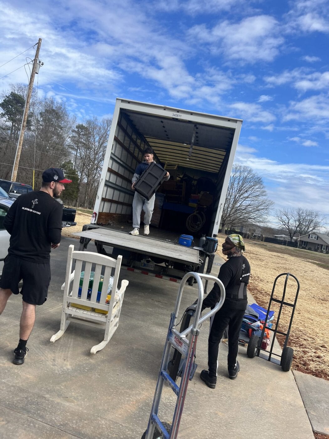 Three people are unloading items from a moving truck onto a driveway, using a dolly for assistance, under a partly cloudy sky.