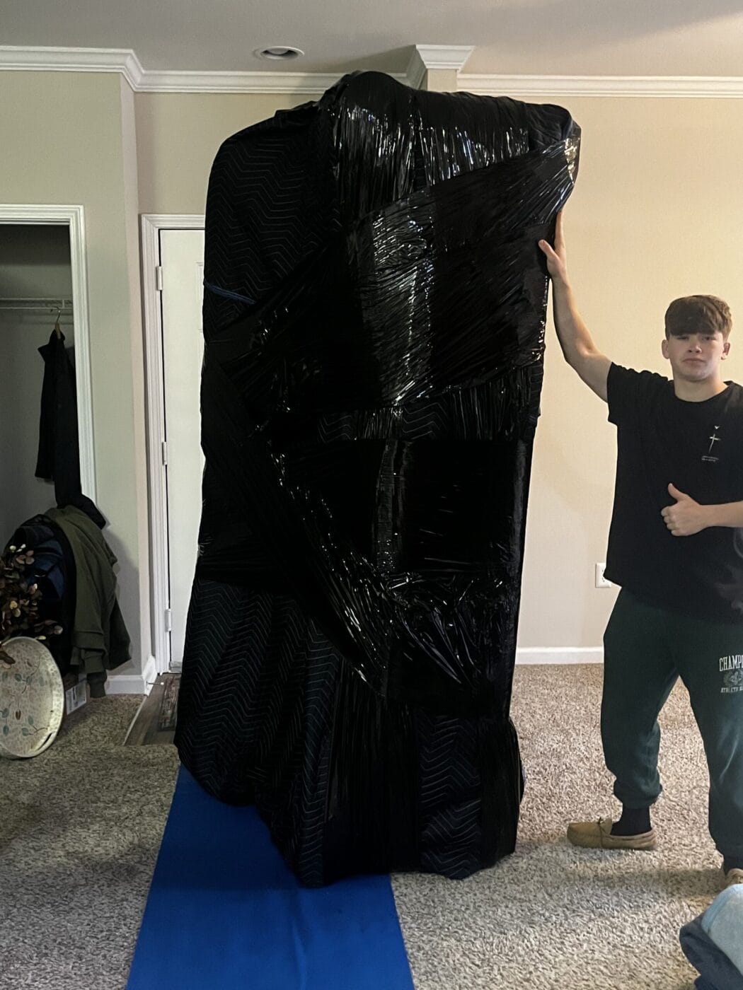 A person stands next to a large object wrapped in black plastic and packing blankets, giving a thumbs-up, showcasing the expertise of White Glove Moving Services.