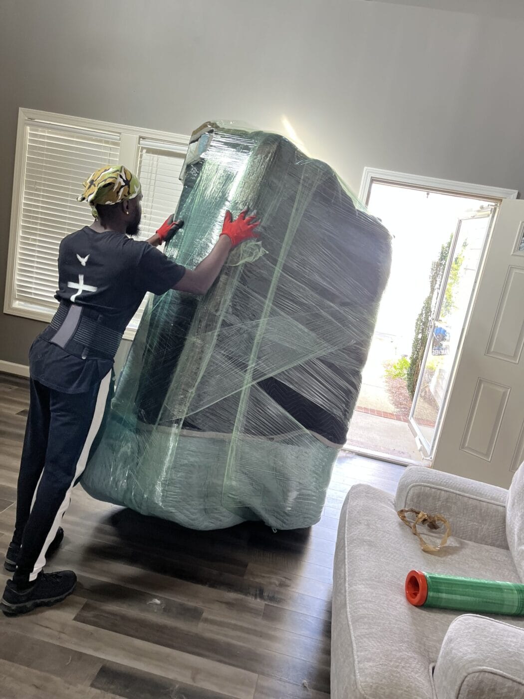 A person wearing a hat and gloves is moving a large, wrapped piece of furniture through a doorway.