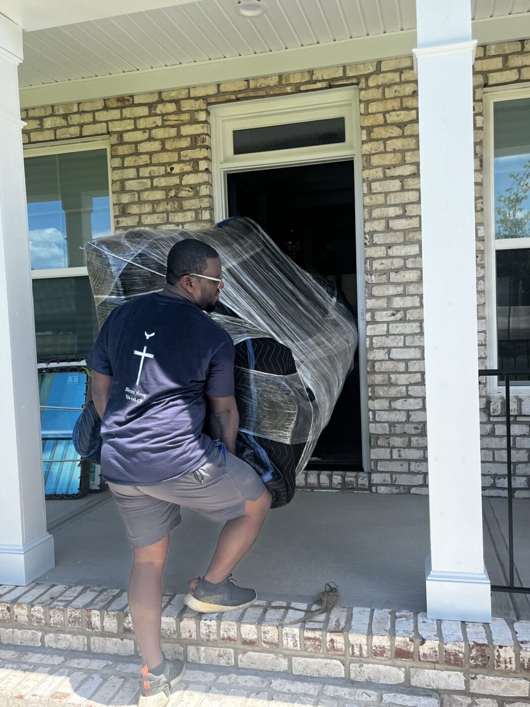 A person wearing a black T-shirt moves a large, wrapped object through a doorway on a porch.